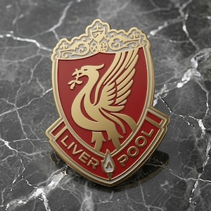 Könnte beinhalten: Eine Metall-Emaille-Anstecknadel mit dem Wappen des Liverpool Football Club. Das Design zeigt einen goldenen Liver-Vogel auf einem roten Schildhintergrund, mit einem dekorativen goldenen Oberteil und einem Banner am unteren Rand mit dem Text LIVER POOL in Gold.