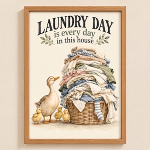 May include: A framed wall art print featuring a large wicker laundry basket overflowing with folded clothing. Beside the basket stands an adult duck with three small ducklings. The text reads: LAUNDRY DAY is every day in this house.