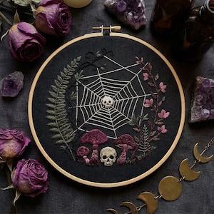 May include: A circular wooden embroidery hoop containing black fabric with white thread stitching of a spider web, a small skull, three purple mushrooms, a second skull, a green fern leaf, and a vine with small pink flowers.
