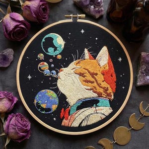 Pode incluir: Um bastidor de bordado circular com fundo de tecido preto apresentando um gato laranja e branco bordado à mão soprando bolhas que lembram planetas coloridos e estrelas. O gato usa uma pequena gravata borboleta vermelha e uma coleira azul-petróleo.