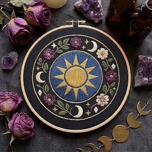 May include: A circular embroidery hoop featuring a central gold sun on a blue background, surrounded by a ring of purple and white flowers, green leaves, and cream-colored crescent moons on a black fabric base, framed in light-colored wood.