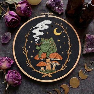 Peut inclure: Un tambour à broder circulaire représentant une grenouille verte assise sur un gros champignon orange, fumant une pipe avec de la fumée blanche. La scène est encadrée par des branches dorées, un croissant de lune jaune et des étoiles sur fond noir.