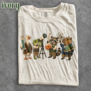 May include: A cream-colored t-shirt featuring a graphic of four animals dressed in vests and bow ties. A duck holds a graduated cylinder, a frog uses a telescope, a raccoon holds a flask, and a capybara holds a globe and atom model.