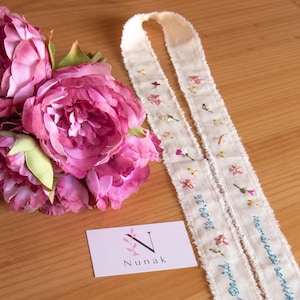 Pode incluir: Um buquê de peônias artificiais rosa-escuro ao lado de uma longa fita de tecido creme com delicados bordados florais e texto azul costurado que diz "Por ti, Belén y David, 26.09.16" e "Momentos siempre". Um pequeno cartão branco exibe o logotipo "N Nunak".