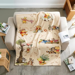 May include: A cream-coloured fleece blanket featuring a repeating pattern of Winnie the Pooh and friends including Tigger, Eeyore, and Piglet. The illustrations show the characters in various poses, such as holding flowers, balloons, and honey pots, scattered across the fabric with small brown leaf motifs.