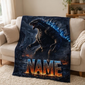 Può includere: Una coperta in pile blu scuro e nero con una grande illustrazione dettagliata di Godzilla con pinne dorsali blu brillante. La creatura si trova in mezzo a un paesaggio urbano in fiamme e in rovina. La parola NAME è stampata in grandi lettere arancioni.
