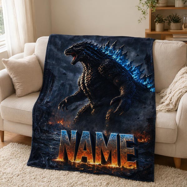 Könnte beinhalten: Eine dunkelblaue und schwarze Fleecedecke mit einer großen, detaillierten Illustration von Godzilla mit leuchtend blauen Rückenflossen. Die Kreatur steht inmitten einer feurigen, zerstörten Stadtlandschaft. Das Wort NAME ist in großen, feurigen Buchstaben gedruckt.