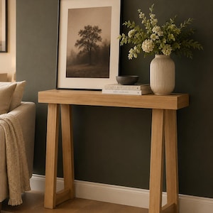 May include: A light oak console table with A-frame legs, topped with a framed sepia-toned tree print, a ribbed cream ceramic vase filled with white flowers, a small dark grey bowl, and two stacked books titled Live Beautifully and This Is Home.