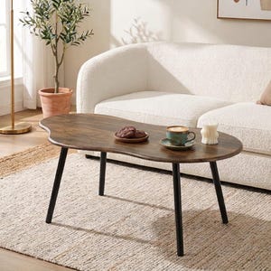 May include: A kidney-shaped coffee table with a dark brown wood grain finish and four slender black metal legs. The tabletop holds a small plate with two chocolate cookies, a teal ceramic coffee cup on a saucer, and a white pillar candle.