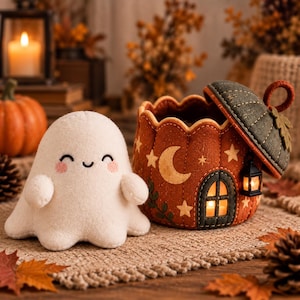 Puede incluir: Un fantasma de peluche blanco con cara sonriente bordada y mejillas rosadas junto a un recipiente de tela naranja redondo con tapa verde oscuro. El recipiente presenta una luna, estrellas, una pequeña puerta arqueada y un farol colgante.