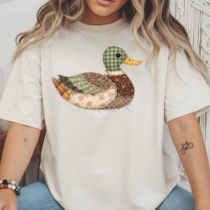 May include: A cream-colored short-sleeve t-shirt featuring a central graphic of a mallard duck. The duck is designed with a patchwork aesthetic, incorporating various fabric patterns including green gingham, brown floral prints, and plaid textures.