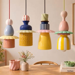 Puede incluir: Cuatro lámparas colgantes con formas de cerámica apiladas en varios colores, incluyendo azul, amarillo, rosa y blanco. Cada luminaria presenta un borde ancho festoneado y está suspendida por un cable fino desde el techo sobre una mesa de madera.