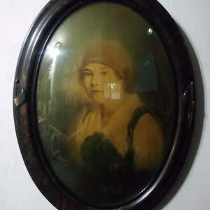 Puede incluir: Retrato ovalado de una mujer con un sombrero rosa claro y una blusa blanca con cuello verde. El retrato está enmarcado en un marco de madera oscura con un diseño de águila en la parte superior.