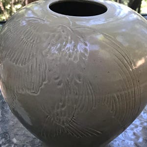 Könnte beinhalten: Eine große, runde, cremefarbene Keramikvase mit einem geprägten Design aus stilisierten Vögeln. Die Vase hat eine breite Öffnung oben.