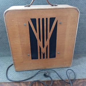 May include: A vintage wooden speaker cabinet with a light brown finish and a dark brown leather handle on top. The front panel features a geometric cutout design revealing a dark fabric speaker grille, secured by four visible metal screws.