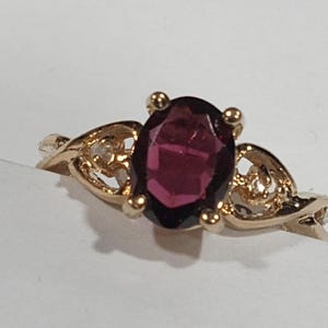 May include: A gold ring with a large, oval-cut, deep red gemstone. The ring has a delicate, filigree design on the band, with small gold accents around the gemstone. The ring is set against a white background.