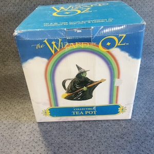 May include: A blue cardboard box for a collectible tea pot featuring The Wizard of Oz branding. The box displays a graphic of a green-skinned witch in a black hat and cloak riding a broomstick, framed by a rainbow. Text includes The Wizard of Oz.