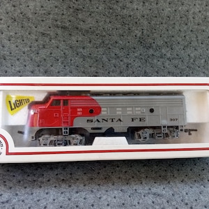 May include: A red and silver Santa Fe HO scale model train locomotive in a white box. The box has the word "Lighted" and the Bachmann logo. The train car has the number 307.
