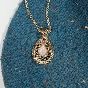May include: A gold-colored pendant necklace with a teardrop-shaped, light-colored stone. The pendant is surrounded by a decorative filigree design and a small pink stone. The necklace chain is also gold-colored.