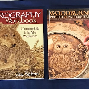 May include: Two instructional books on woodburning. The left book is titled Pyrography Workbook: A Complete Guide to the Art of Woodburning by Sue Walters, featuring a wolf illustration. The right book is Woodburning Project & Pattern Treasury.