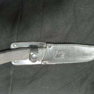 May include: A black knife with a textured handle is sheathed in a black leather case. The case has a metal clasp and the brand name "CRKT" is visible. The knife is designed for outdoor use.