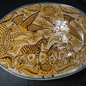 May include: A white oval ceramic plate with a blue rim. The plate is decorated with a yellow and black floral design featuring birds and zebras.