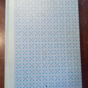 May include: A hardcover book with a light blue spine and a patterned cover. The cover features a repeating floral design in shades of blue and purple on a white background. The book is closed and appears to be in good condition.