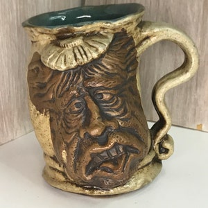May include: A ceramic mug with a brown, textured face and a light brown handle. The mug has a light brown and white glaze with a blue rim.