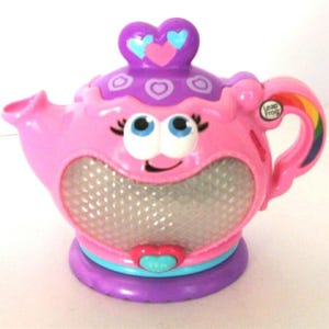 Puede incluir: Una tetera de juguete de plástico rosa con una tapa morada con diseños de corazones. La tetera tiene una cara sonriente con ojos azules, un panel frontal plateado texturizado, un asa de arcoíris y un botón en forma de corazón con LeapFrog.