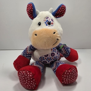 Puede incluir: Un juguete de peluche con forma de vaca que presenta una cara blanca con hocico beige, un parche en el ojo con estrellas y orejas con lunares rojos y blancos. El cuerpo está cubierto de tela con motivos rojos, blancos y azules.