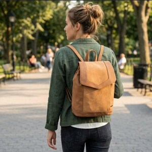 Peut inclure: Un sac à dos en daim beige doté d'un rabat, d'une poignée supérieure et de bretelles réglables. Le sac présente une conception structurée avec un compartiment principal rectangulaire et une petite étiquette rectangulaire sur le côté.
