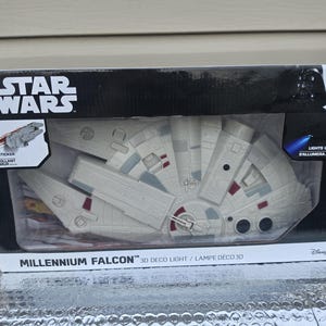 May include: A Star Wars Millennium Falcon 3D deco light in its original packaging. The box features the Star Wars logo, a graphic of a wall sticker, and text indicating it lights up. The light is a white, grey, and red plastic model of the ship.