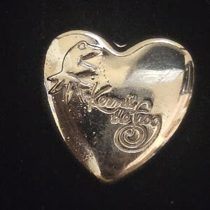 Puede incluir: Un broche de metal dorado en forma de corazón con un gráfico en relieve de una cabeza de rana y el texto Kermit the Frog en letra cursiva. La superficie del broche es lisa y reflectante con un acabado pulido y detalles grabados claros.
