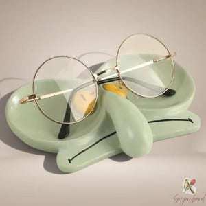 Puede incluir: Un soporte para gafas de cerámica verde claro con forma de cara y una nariz prominente, con una línea negra que representa la boca. Sobre el soporte descansan unas gafas redondas de montura dorada, lentes transparentes y patillas negras.