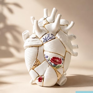 May include: A white ceramic anatomical heart sculpture featuring a kintsugi-style design with gold-colored seams connecting various porcelain fragments. Some segments display floral patterns in red, blue, and green, while others are plain white.