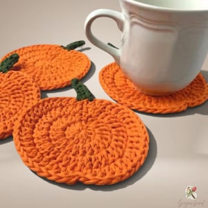 Puede incluir: Tres posavasos de ganchillo de color naranja con forma de calabaza, cada uno con un pequeño tallo verde oscuro en la parte superior. Un posavasos sostiene una taza de cerámica blanca, mientras que los otros dos están colocados cerca.