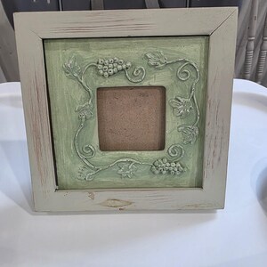 Puede incluir: Un marco de fotos cuadrado con un borde de madera de color beige claro desgastado. El panel interior está pintado en un suave verde salvia y presenta un relieve moldeado de racimos de uvas y vides que rodean la abertura central.