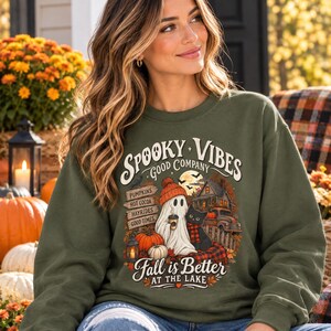 May include: An olive green crewneck sweatshirt featuring a graphic print with the text SPOOKY VIBES, GOOD COMPANY, and Fall is Better AT THE LAKE. The design includes a ghost in an orange beanie, a black cat, pumpkins, and a rustic cabin.