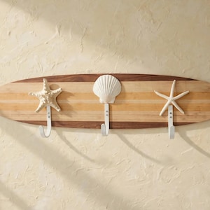 May include: A wall-mounted coat rack shaped like a surfboard made of light and dark wood. It features three white metal hooks, each topped with a decorative sea element: a beige starfish, a white scallop shell, and a white starfish.