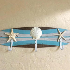 May include: A wall-mounted coat rack shaped like a surfboard with a distressed finish in brown, white, and turquoise stripes. It features three white metal hooks, each adorned with a natural sea element: a starfish, a scallop shell, and a starfish.
