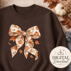 May include: A dark brown crewneck sweatshirt featuring a central graphic of a large, cream-coloured ribbon tied into a bow. The ribbon is decorated with a repeating pattern of small orange pumpkins and brown autumn leaves, with loose leaves scattered around.