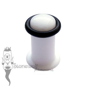 May include: A white ear plug with a smooth, rounded top surface and a black O-ring encircling the upper edge. The body of the plug is cylindrical with a flared base, and the text Stonewolf Adornments is printed in black near the bottom left.