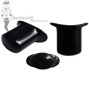 Può includere: Tre dilatatori per orecchie in acrilico nero di diverse dimensioni e forme. Il dilatatore più grande è un cilindro con una parte superiore e inferiore svasata. Il dilatatore medio è un cilindro con una parte superiore svasata. Il dilatatore più piccolo è un ovale piatto.