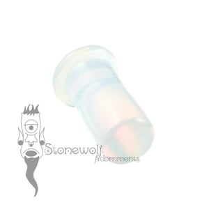 May include: A translucent, cylindrical ear plug with a flared end, featuring a smooth, semi-clear finish that catches the light. The object is positioned diagonally, accompanied by a logo featuring a stylized cyclops head and the text Stonewolf Adornments.