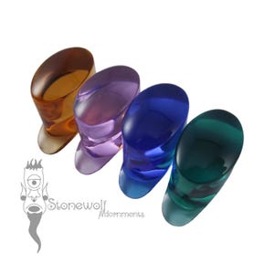 May include: Four translucent, oval-shaped ear plugs in amber, purple, blue, and green. Each piece features a smooth, polished finish and a flared end. A logo with a cyclops graphic and the text Stonewolf Adornments is visible in the corner.