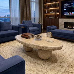 May include: A large, oval-shaped coffee table made of light beige, textured stone with a rough, natural edge. The table is supported by two thick, rounded stone pedestals and sits in a living room with blue velvet furniture and a fireplace.