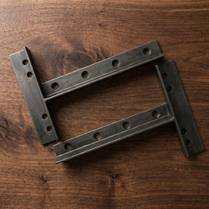 May include: Two L-shaped metal shelf brackets with a dark, weathered finish. Each bracket features a series of pre-drilled circular holes along both the vertical and horizontal segments, designed for secure mounting to a wall and a shelf surface.