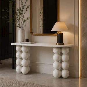 Può includere: Un tavolo console bianco con un piano liscio e due gambe composte da sfere bianche impilate. Sul tavolo si trovano un alto vaso cilindrico bianco con rami, un piccolo oggetto marrone scuro, un libro e una lampada nera con paralume bianco.
