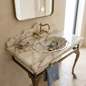 May include: A white marble vanity with brown veining features a scalloped basin, a brass faucet, and a matching brass stand with curved legs. A small soap dish with a bar of soap, a seashell, and a blue waffle-weave towel are present.