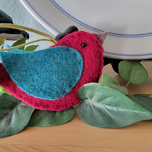 Może przedstawiać: Filcowa ozdoba w kształcie ptaka z czerwonym ciałem, niebieskimi skrzydłami i czarnym okiem. Ptak siedzi na łóżku z zielonych liści.